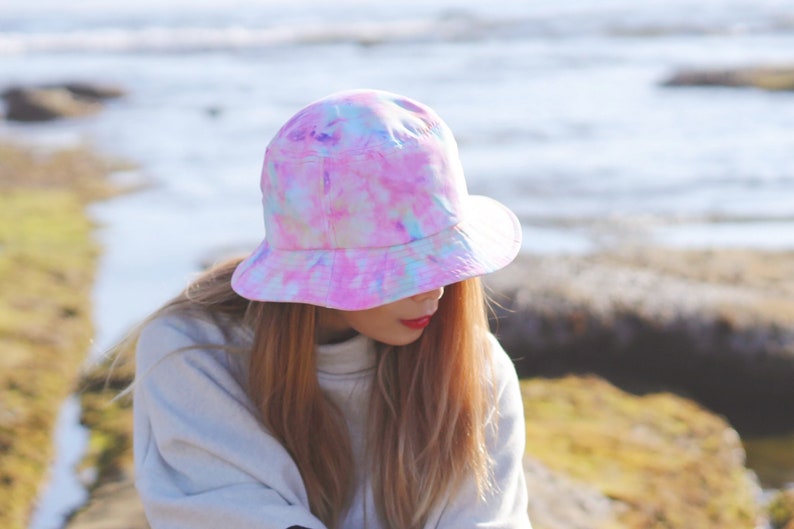 May include: A person wearing a pink, blue, and white tie-dye bucket hat. The hat has a wide brim and is made of a soft, comfortable fabric.