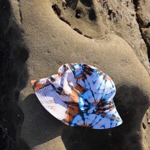 May include: A white bucket hat with a tie-dye pattern in shades of blue, orange, and brown. The hat is lying on a sandy surface.