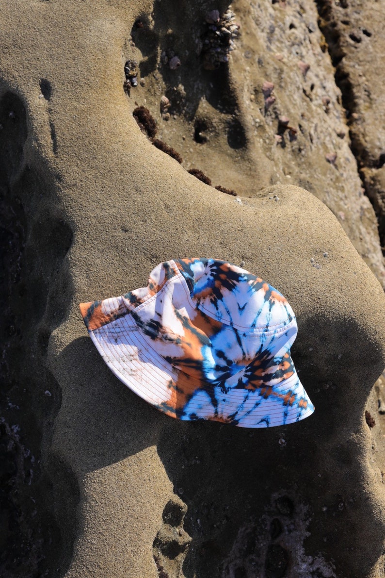 May include: A white bucket hat with a tie-dye pattern in shades of blue, orange, and brown. The hat is lying on a sandy surface.