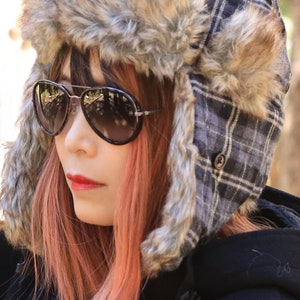 May include: A woman with pink hair is wearing a black jacket and a gray and white plaid trapper hat with a brown fur trim. She is wearing aviator sunglasses.