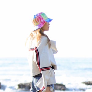 May include: A woman wearing a white jacket with red and blue stripes, blue denim shorts, and a tie-dye bucket hat. She is standing on a beach with the ocean in the background.