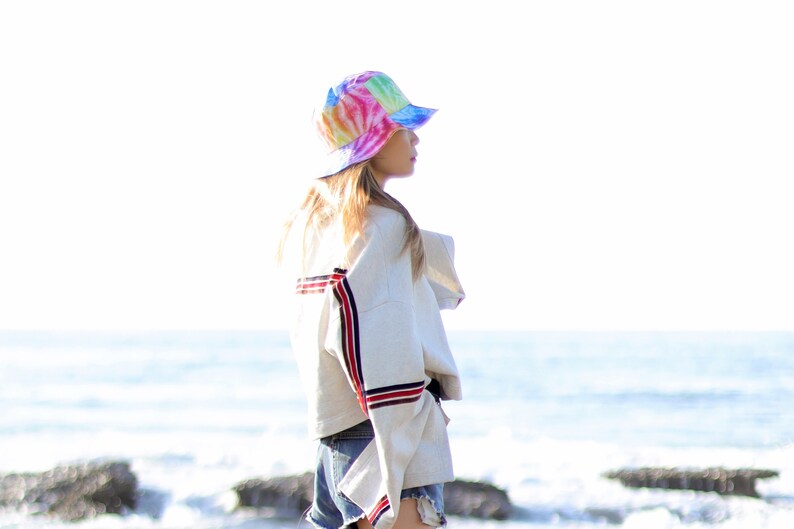 May include: A woman wearing a white jacket with red and blue stripes, blue denim shorts, and a tie-dye bucket hat. She is standing on a beach with the ocean in the background.