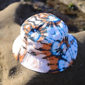 May include: A white bucket hat with a tie-dye pattern in shades of blue, orange, and black. The hat is lying on a sandy surface.