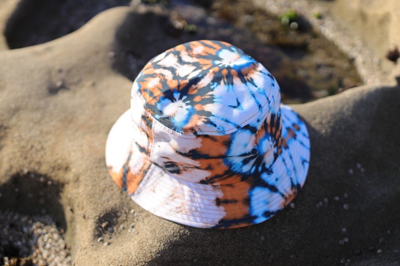 May include: A white bucket hat with a tie-dye pattern in shades of blue, orange, and black. The hat is lying on a sandy surface.