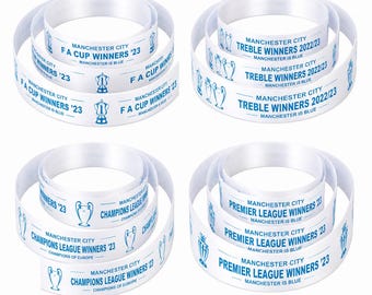 May include: Four white ribbons with text and blue graphics. Each ribbon is coiled, displaying phrases like "FA Cup Winners '23" and "Treble Winners 2022/23". The text "Manchester City" and "Manchester is Blue" are also visible.