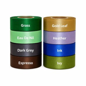 May include: A collection of ribbon spools in various colors, including green, gold, purple, and blue. Each spool is labeled with its color name: Grass, Gold Leaf, Heather, Ink, and Ivy. The ribbons are neatly stacked, showcasing their vibrant hues.