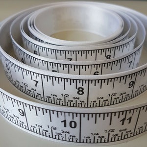May include: White measuring tape with black markings in inches and fractions of an inch. The tape is rolled up and shows numbers 6 through 11.