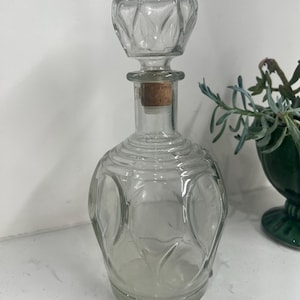 Puede incluir: Una decantador de vidrio transparente con un diseño texturizado y facetado. La decantador tiene un tapón de corcho y una parte superior redonda y facetada.