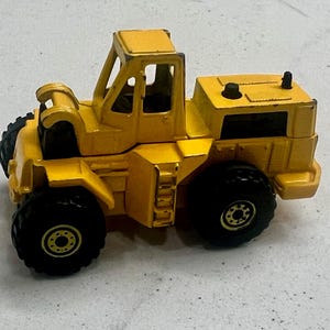 Puede incluir: Un juguete amarillo de cargadora frontal con neum&aacute;ticos negros. El juguete tiene una cabina con una barra antivuelco y un compartimento del motor trasero. La cargadora es una r&eacute;plica en miniatura de un veh&iacute;culo de construcci&oacute;n.