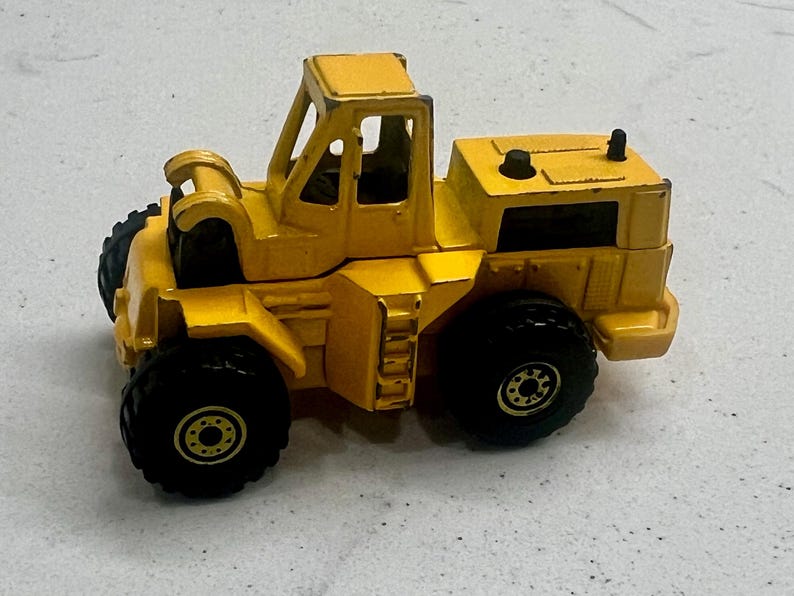 Puede incluir: Un juguete amarillo de cargadora frontal con neum&aacute;ticos negros. El juguete tiene una cabina con una barra antivuelco y un compartimento del motor trasero. La cargadora es una r&eacute;plica en miniatura de un veh&iacute;culo de construcci&oacute;n.