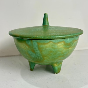 May include: A green ceramic bowl with three short legs and a matching lid featuring a central pointed handle. The surface displays a mottled texture with blended shades of light and dark green, creating a variegated appearance across the exterior.