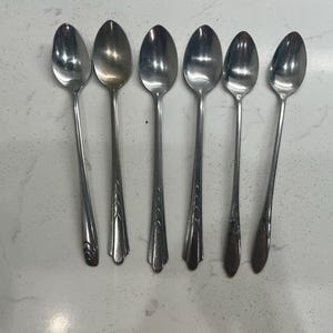 May include: Six vintage silver spoons on a white surface. The spoons have elongated bowls and decorative handles with different patterns. The spoons are made of a shiny, reflective metal.