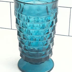 May include: A teal-colored, vintage-style drinking glass. The glass has a textured, geometric design around the body and a circular base. The glass is approximately 6 inches tall.