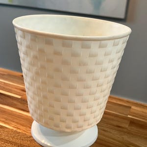 May include: Off-white, woven-patterned planter with a wide, open top. The planter sits on a white, circular base. The textured design gives the planter a basket-like appearance. The planter is suitable for indoor or outdoor use.