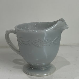 Könnte beinhalten: Ein grauer Glas-Sahnegießer mit einem Blattdesign um den oberen Rand. Der Sahnegießer hat einen Griff und einen runden Boden.