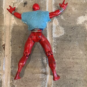 以下が含まれることがあります： 赤いアクションフィギュアが青いシャツを着て仰向けに寝ています。フィギュアは腕と脚が可動式です。