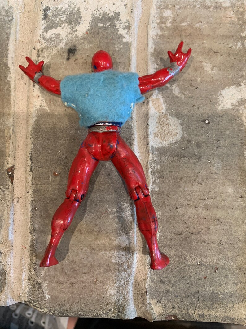以下が含まれることがあります： 赤いアクションフィギュアが青いシャツを着て仰向けに寝ています。フィギュアは腕と脚が可動式です。