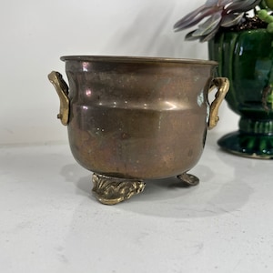 Puede incluir: Una pequeña olla de latón antigua con dos asas y tres patas decorativas. La olla tiene un cuerpo redondeado y un borde ligeramente ensanchado. El latón tiene una pátina cálida y envejecida, lo que sugiere su antigüedad. Una pieza decorativa encantadora.