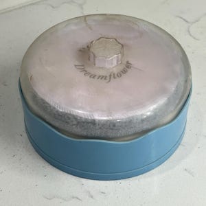May include: A vintage powder compact with a clear, domed lid and a light blue base. The lid has a flower-shaped knob and the words "Dreamflower" are printed on the lid. The compact contains a powder puff.