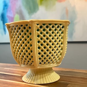 Puede incluir: Una jardinera de color amarillo pálido con un diseño de celosía y una base de pedestal. El interior está forrado con material verde. La jardinera está hecha de un material duradero y es adecuada para uso en interiores o exteriores. Es un artículo decorativo.