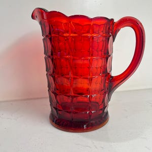 May include: A vibrant red glass pitcher featuring a prominent grid-like embossed pattern across its exterior surface. The vessel includes a curved handle on the side, a scalloped rim, and a small pouring spout at the top, showcasing a translucent finish.