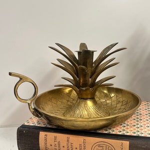 May include: A gold pineapple-shaped candle holder with a small dish for wax. The holder has a handle and a detailed pineapple design.