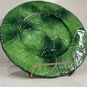 Pu&ograve; includere: Un grande piatto in ceramica verde con un motivo a foglie. Il piatto &egrave; su un supporto in metallo dorato.