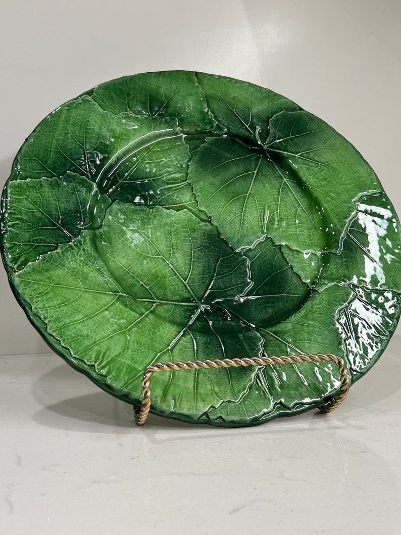 Pu&ograve; includere: Un grande piatto in ceramica verde con un motivo a foglie. Il piatto &egrave; su un supporto in metallo dorato.
