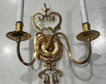 May include: A gold-toned, ornate, two-arm wall sconce with a decorative scroll design. The sconce has two candle holders with white candles.