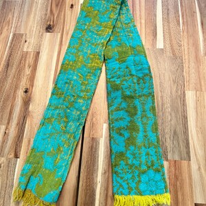 May include: A long, rectangular scarf with a vibrant pattern of teal and olive green. The scarf has a textured appearance and is finished with yellow fringe at both ends. The design features a floral or abstract motif.