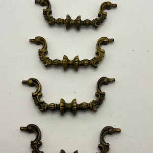 Può includere: Quattro tiretti per cassetti in ottone antico con disegni decorativi. Ogni tiretto ha una forma curva con un motivo a volute decorative e un pomo centrale.