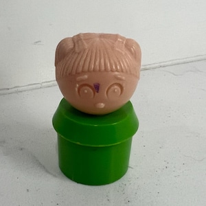Könnte beinhalten: Eine Vintage-Spielfigur mit einem hellpfirsichfarbenen Kopf, geformten Haaren und Gesichtszügen, auf einem hellgrünen zylindrischen Sockel. Das Spielzeug hat ein einfaches, klassisches Design, wahrscheinlich aus einem Kinderspielzeug-Set.