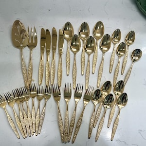 Puede incluir: Una colección de cubiertos dorados, que incluye tenedores, cuchillos, cucharas y utensilios para servir. Los utensilios tienen un patrón floral en los mangos. La disposición está sobre una superficie blanca.