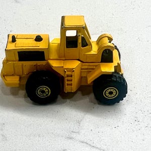 Puede incluir: Un juguete amarillo de cargadora frontal con neum&aacute;ticos negros. El juguete tiene una cabina con una escalera y un compartimento trasero. Los neum&aacute;ticos tienen centros dorados. El juguete es de metal y pl&aacute;stico.