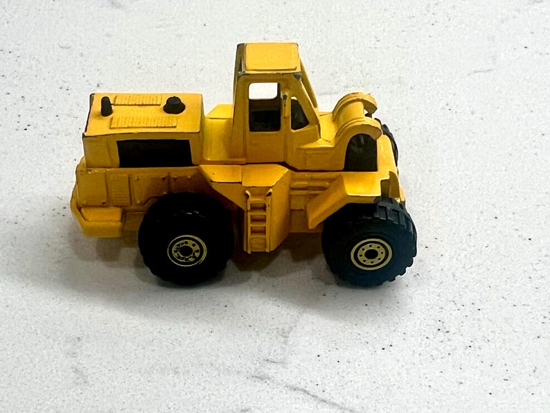 Puede incluir: Un juguete amarillo de cargadora frontal con neum&aacute;ticos negros. El juguete tiene una cabina con una escalera y un compartimento trasero. Los neum&aacute;ticos tienen centros dorados. El juguete es de metal y pl&aacute;stico.