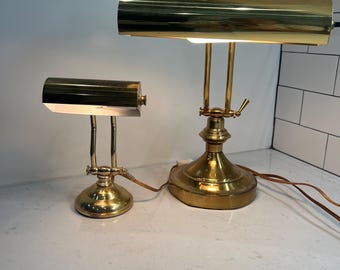 May include: Two polished brass piano lamps of different sizes, each featuring a cylindrical shade, a dual-post arm, and a circular base. Both lamps have a metallic gold finish and are connected to brown electrical cords, with the larger lamp positioned to the right of the smaller one.