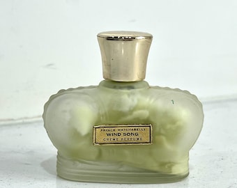 Puede incluir: Un frasco de perfume crema Wind Song de Prince Matchabelli vintage. El frasco tiene un tono verde claro esmerilado con una tapa dorada. La etiqueta dice "Prince Matchabelli Wind Song Creme Perfume". El frasco tiene una forma única de corona.