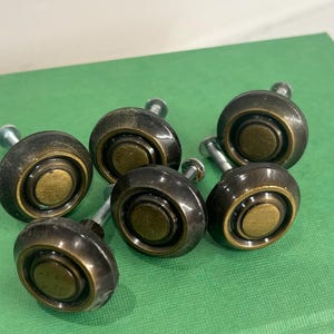May include: Six antique-style cabinet knobs with a dark bronze finish and brass accents. Each knob has a rounded shape with a central brass disc and a threaded post for mounting. The knobs are arranged on a green surface.