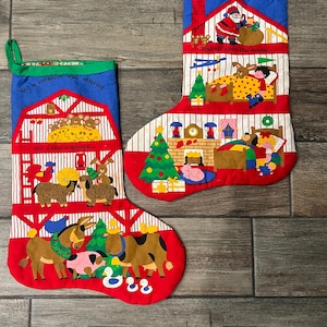 May include: Two fabric Christmas stockings featuring colorful holiday illustrations. The left stocking displays a barn scene with animals and the text 'Not a creature was stirring, not even a mouse'. The right stocking shows a house interior with Santa and the text 'Twas the night before Christmas and all thru the house'.