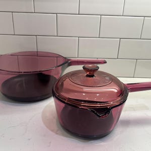 May include: Two translucent purple glass saucepans with long handles. One saucepan is open, while the other features a matching glass lid with a circular knob handle and a textured sunburst pattern on the top surface of the lid.