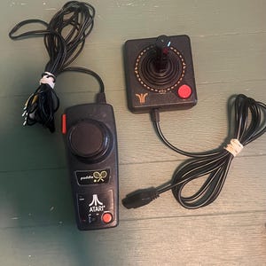 May include: Two vintage Atari video game controllers. One is a joystick with a black base, a red button, and a black joystick. The other is a paddle controller with a black base, a red button, and a dial. Both have black cords.