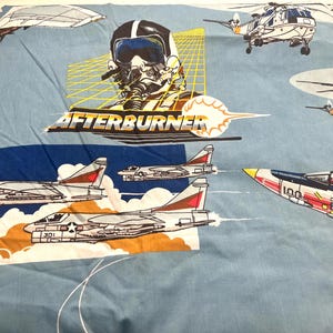 Puede incluir: Sábana azul claro con un gráfico de piloto de combate y la palabra "AFTERBURNER". La sábana presenta ilustraciones de aviones de combate, helicópteros y otras aeronaves. El diseño incluye un esquema de color azul y blanco.