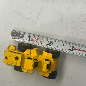 Puede incluir: Un veh&iacute;culo de construcci&oacute;n de juguete amarillo con ruedas negras, de aproximadamente 7,6 cm de largo, est&aacute; colocado junto a una cinta m&eacute;trica. La cinta, marcada en pulgadas, indica el tama&ntilde;o del veh&iacute;culo. La cinta est&aacute; marcada "Made in USA."