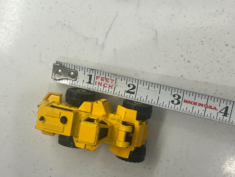 Puede incluir: Un veh&iacute;culo de construcci&oacute;n de juguete amarillo con ruedas negras, de aproximadamente 7,6 cm de largo, est&aacute; colocado junto a una cinta m&eacute;trica. La cinta, marcada en pulgadas, indica el tama&ntilde;o del veh&iacute;culo. La cinta est&aacute; marcada "Made in USA."