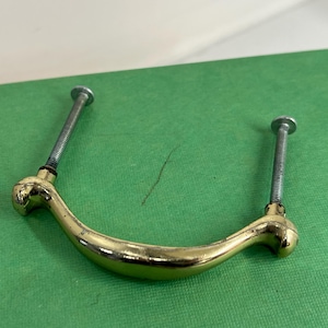 May include: A gold-colored, curved drawer pull with two silver screws. The pull has a rounded design and is set against a green background. The screws are threaded and have flat heads.