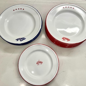 Puede incluir: Platos de esmalte blanco con detalles en rojo, blanco y azul. Los platos tienen un borde rojo o azul y presentan un diseño de vagón rojo o avión azul. Algunos platos tienen estrellas rojas.