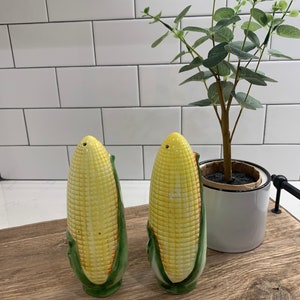 May include: Two ceramic corn on the cob salt and pepper shakers. The shakers are yellow with green husks and are sitting on a wooden tray. A small potted plant is behind the shakers.