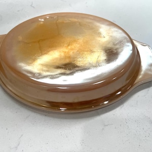 May include: An iridescent, oval-shaped casserole dish with two handles. The dish has a rounded lid and a warm, amber-colored finish. The dish is likely made of glass and is suitable for baking and serving.