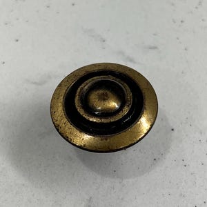 Puede incluir: Pomo de armario redondo de latón antiguo con diseño de círculos concéntricos. El pomo tiene un aspecto ligeramente envejecido, con un centro oscuro y un borde dorado-bronce. Adecuado para la restauración de muebles o para añadir encanto vintage.