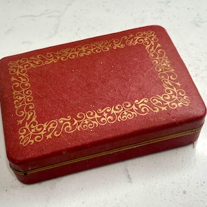 May include: A rectangular red box with a gold decorative border. The box has a textured surface and a gold trim. The gold border features an ornate, swirling design. The box is closed and appears to be made of leather or a similar material.